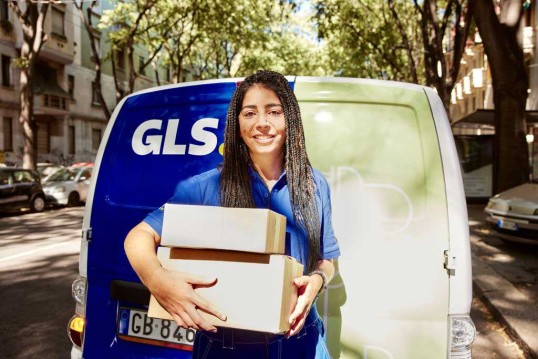 • Repartidora de GLS sosteniendo paquetes de mensajería para entrega de última milla. Servicio de transporte de paquetería eficiente y logística urbana con furgoneta sostenible.