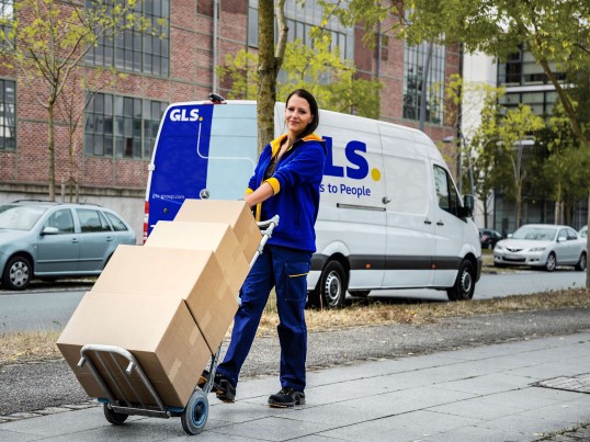 GLS delivery woman transporting multiple parcels on a hand truck, with a GLS delivery van in the background.